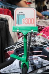 Placa de Promo&ccedil;&atilde;o com Valor 300 em Feira de Moda Circular ao Ar Livre, Economia Circular e Roupas Second-Hand, Detalhe de Bra&ccedil;o de Pessoa Selecionando Roupa ao Fundo