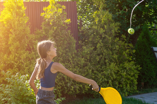 Energetic pre-teen girl playing with yellow tennis racket and tethered ball in sun-drenched green garden during vibrant summer day