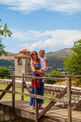 Mother and daughter pose for heartwarming portrait on rustic wooden bridge, overlooking historic stone tower and picturesque mountain village under bright summer sky