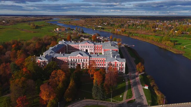 Aerial pans over Jelgava Palace in Jelgava, Latvia, by the Lielupe River. The U shaped palace, promenade, bridge, cars, fields, and rooftops appear in crisp fall light.