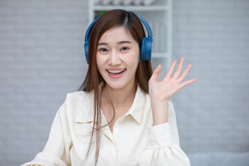 Asian smiling woman wearing blue headphones waving at camera with friendly expression in bright indoor setting, long hair and white blouse creating casual modern portrait