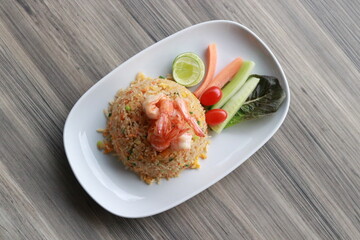 A plate of delicious shrimp fried rice with fresh vegetable garnishes. The dish is presented on a white plate and placed on a wooden table.