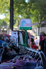 Feira de Moda Circular ao Ar Livre com Roupas em Araras, Clientes Desfocados ao Fundo e Placa de Promo&ccedil;&atilde;o Destacando Valor 300, Ambiente Natural