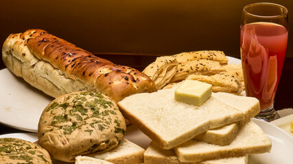 A warm and appetizing food composition featuring assorted bakery items including freshly baked bread slices, buns topped with herbs, puff sticks, butter, and a glass of fruit juice