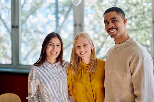Three diverse young colleagues standing together in a modern office, happily smiling and representing teamwork and partnership