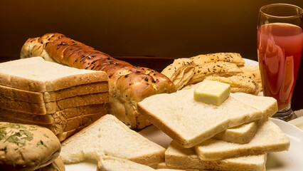 A warm and appetizing food setup featuring slices of white bread topped with butter, a loaf of garlic bread, puff sticks, and a glass of red fruit juice on a golden background