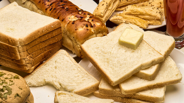 A close-up view of assorted bakery breads, white toast slices with butter, puff sticks, and a loaf of garlic bread arranged on a plate. The image captures the freshness and warmth