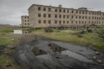 Crumbling Russian apartment blocks with shattered windows and overgrown grass in Ugolnye Kopi, Chukotka, Russia, under cloudy summer skies.
