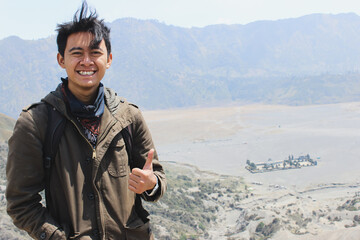 Smiling Young Asian Traveler Giving Thumbs Up at Mount Bromo Volcano Landscape in Indonesia. symbolizing approval, success, and a great journey.