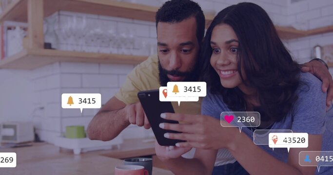 Leaning couple viewing black tablet in modern kitchen, with coffee mugs and notification icons