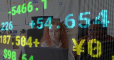 Two colleagues wearing office shirts leaning over laptop screen at office desk, with stock tickers