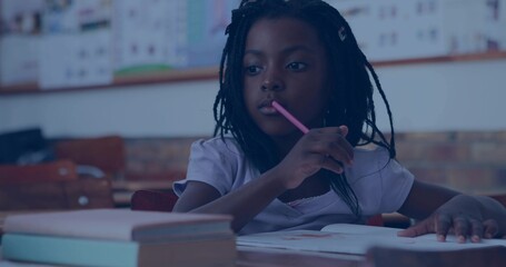 Sitting girl holding pink pencil near mouth, looking upward at open notebook in school classroom
