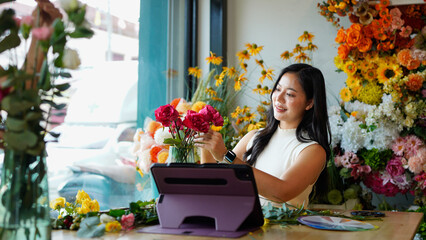 A woman is sitting at a table with a laptop and a vase of flowers in front of her. She is smiling...