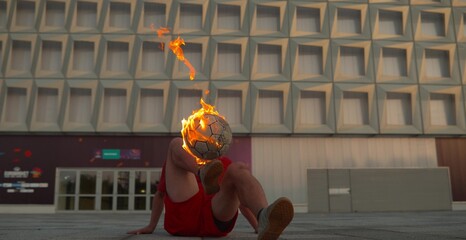 Freestyle football player performing trick with burning soccer ball outdoors in urban area at sunset, concept of extreme sports and creative street performance