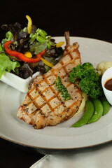 A close-up shot of a gourmet grilled pork chop with various side dishes on a plate. The chop is nicely grilled with grill marks, with a side salad and some green beans.