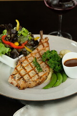 Delicious grilled pork chop served with a fresh salad and a glass of red wine. The pork chop is placed on a white plate with side dishes