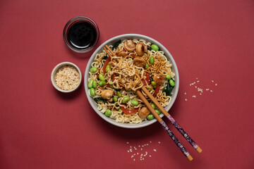Asian noodle bowl with edamame beans, mushrooms and vegetables on dark red background, minimalist top view of healthy vegan meal