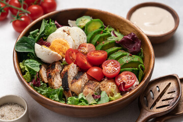Healthy Cobb salad with grilled chicken breast, avocado, cherry tomatoes, lettuce and boiled egg in wooden bowl, top view on light background