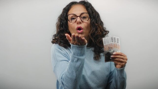 Woman holding icelandic krona banknotes blows a kiss to the camera against a white wall background expressing joy.