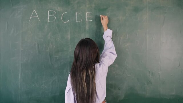 Video English basic class online concept. Happy young Asian female teacher stands writing and teaching English language lesson on the blackboard via video call on a technology smartphone in classroom.