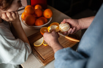 Close-up of man's hands peeling fresh orange on wooden cutting board while child watching. Bowl filled with whole oranges on kitchen background.  Homemade orange   juice preparation, family life.