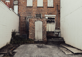 An old brick building with boarded windows and a weathered wooden door sits between two white walls weeds grow by the entrance and cracked pavement leads to the steps