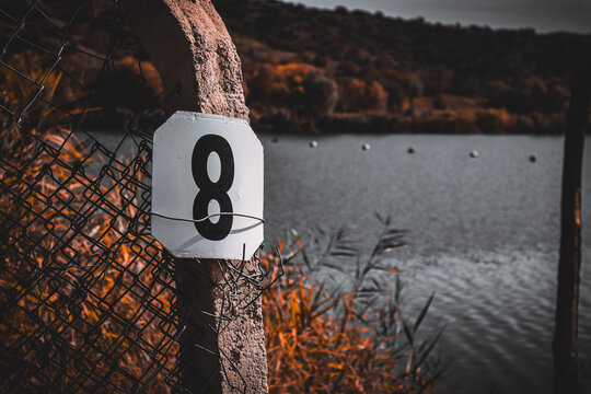 Number eight sign on a concrete post with rusty fence wire and lake background in autumn colors.