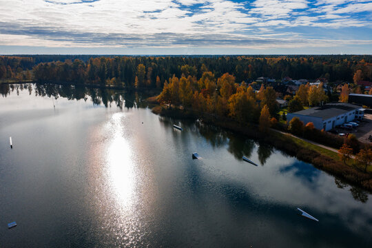 Aerial view of a lake in Jelgava, Latvia with forest, small settlement, docks, and floating obstacles for water sports, in low autumn sun with long shadows and high clouds.