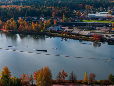Aerial view over Jelgava, Latvia, shows a calm river, autumn trees, and a wakeboarding park with obstacles. Low industrial buildings and houses line the shore in warm light.
