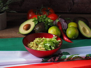 Guacamole in a red bowl, with ingredients at the back. All displayed over a Mexican flag.