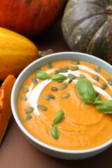 Delicious pumpkin soup with seeds in bowl, basil and vegetables on brown background, closeup