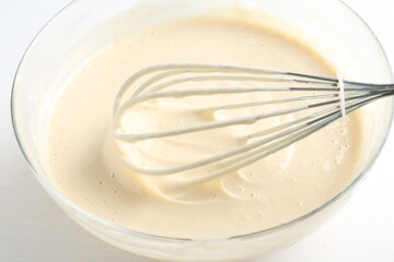 Bowl and whisk with batter (liquid dough) on white table, closeup