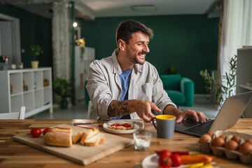 Man smiling working on laptop having breakfast at home