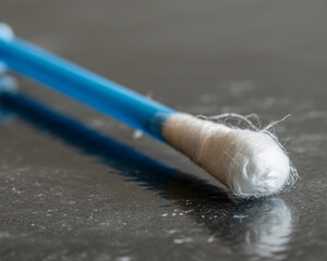 Close up of a clean cotton swab with a blue plastic handle resting on a dark textured surface, highlighting its delicate fibers and essential utility