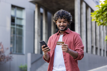 Young Indian man standing outside, looking at phone screen and using credit card