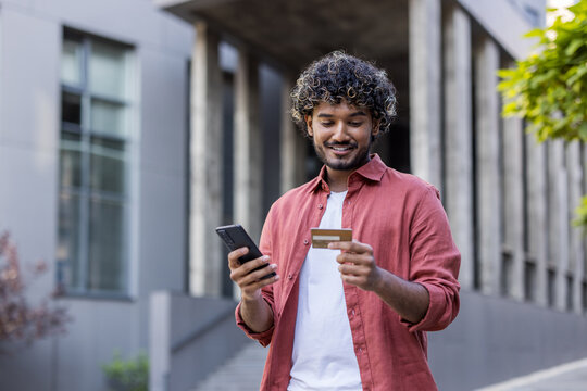 Young Indian man standing outside, looking at phone screen and using credit card