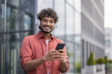 Portrait of a young Indian man wearing white headphones holding a phone and smiling at the camera
