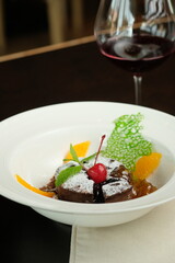 A beautifully presented chocolate lava cake with a glass of red wine. The dessert is garnished with a cherry and orange slices and in a bowl, a glass of red wine in the background