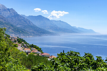 Scenic view of the Croatian coast with blue Adriatic Sea, green vegetation, red-roofed houses, and majestic mountains rising in the background under a clear summer sky.