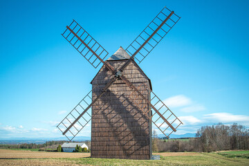 Historic wooden windmill standing in a rural field under a clear blue sky, with distant mountains and farm buildings in the background. Traditional countryside scenery.