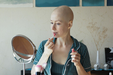Smiling woman with bald head applies lip gloss at home, completing her makeup routine after...