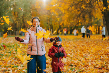 A woman and child joyfully play in a park amidst falling autumn leaves, surrounded by golden hues.