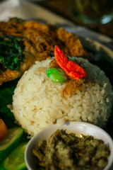 Traditional Indonesian Nasi Liwet served on a wooden table. The dish includes steamed rice, fried tempeh or egg topping, vegetables, sambal, and herbal tea.