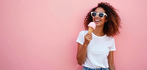 Happy young woman with afro hairstyle eats pink ice cream in waffle cone. Cheerful smiling girl wearing sunglasses holds sweet frozen dessert. Female model isolated on pink background laughing,
