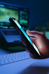 Close-up of a hand holds smartphone and scrolling through social media in dark room, symbolizing late-night content consuming, doom scrolling and digital addiction habits. 