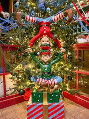 Colorful holiday decorations featuring three cheerful elves stacked together in a playful pose, surrounded by a beautifully adorned Christmas tree with twinkling lights