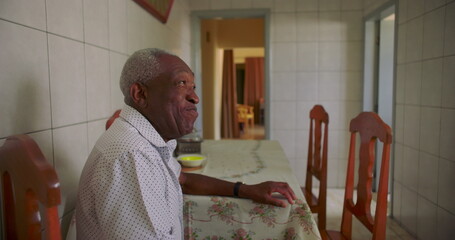 Elderly African American man smiling warmly while seated at a dining table in a cozy kitchen,...
