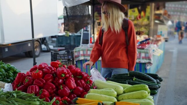 Woman with backpack browsing lively outdoor farmers market stall, selecting vibrant red bell pepper among fresh organic produce for healthy meal