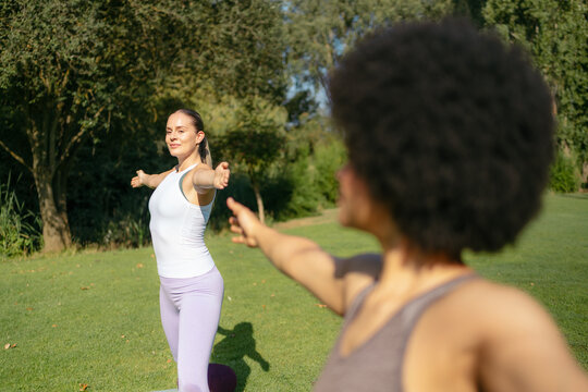 Two diverse women performing yoga postures together in a sunny park with trees