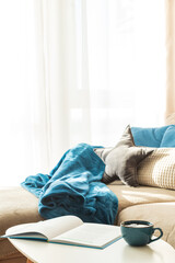 Cozy Living Room with mug and book on Coffee Table, Cushions, Blanket and Sunlight on Sofa © thayra83